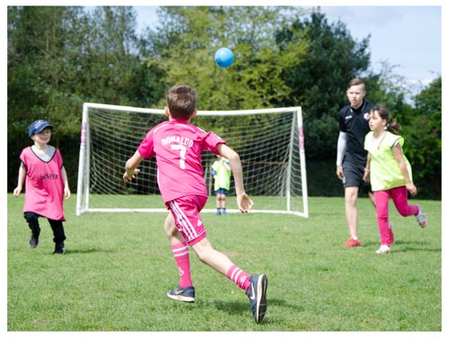 a child kicking a football towards a goal while a footiebugs coach watches on