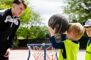 child putting a ball in a basketball hoop while a bugs group coach watches