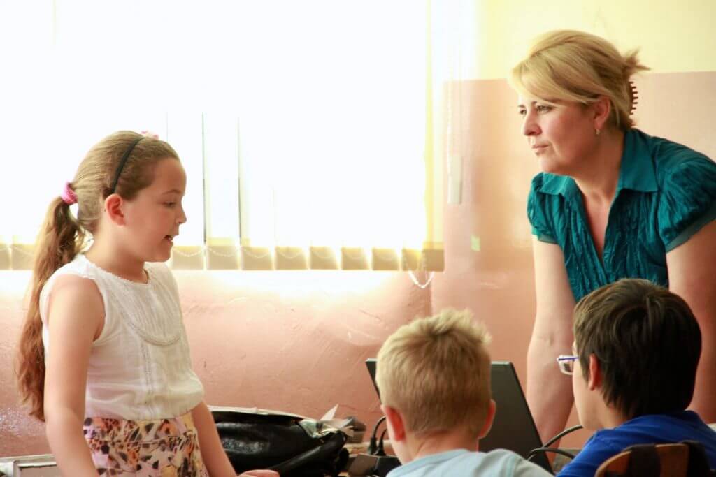 a child talking in front of two other children while an adult watches on