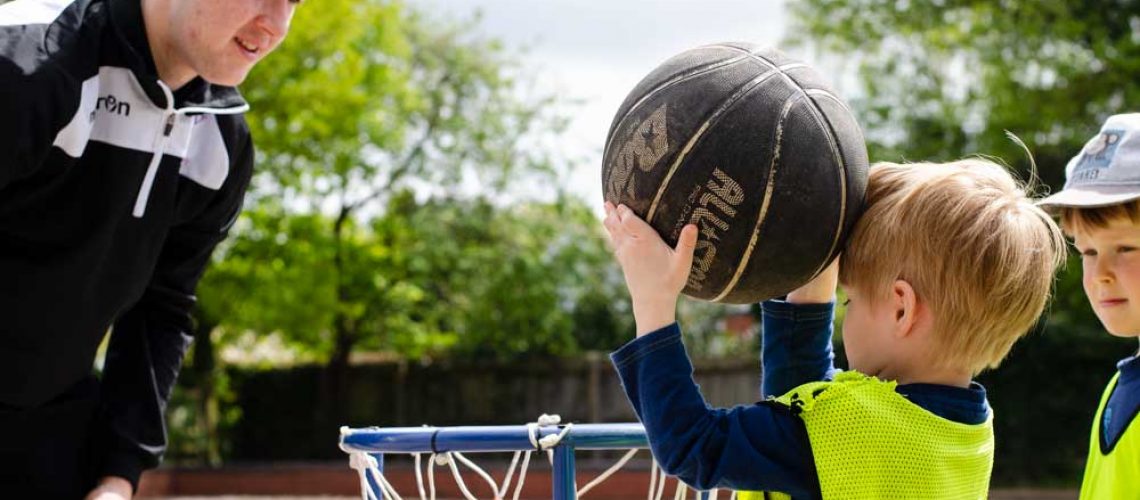 child putting a ball in a basketball hoop while a bugs group coach watches